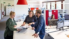 Drei Personen stehen sprechend an einem Tisch