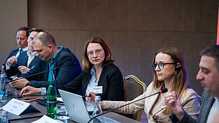 Fünf Personen sitzen an einem Konferenztisch mit Mikrofonen und Laptops, eine Person trägt Kopfhörer