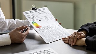 Zwei Personen am Tisch, eine zeigt mit Stift auf ein CO2-Emissionslabel für Pkw mit farbiger Skala von grün bis rot