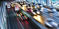 Bewegte Autos auf einer mehrspurigen Straße, mit verschwommenen Lichtern und reflektierenden Oberflächen. Die Straße verläuft unter einer Brücke.