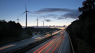 Dämmerungsaufnahme einer Autobahn mit fließendem Verkehr. Im Hintergrund stehen mehrere Windkraftanlagen und der Himmel ist bewölkt.