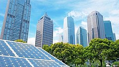 Solaranlage im Vordergrund mit blauen Solarpanelen, im Hintergrund moderne Wolkenkratzer und grüne Bäume unter blauem Himmel.