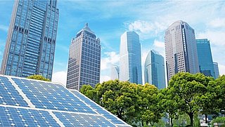 Solaranlage im Vordergrund mit blauen Solarpanelen, im Hintergrund moderne Wolkenkratzer und grüne Bäume unter blauem Himmel.