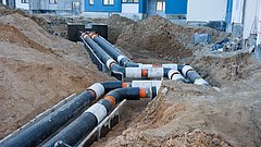 Große schwarze Rohrleitungen mit weißen Isolierungen verlaufen durch eine Baustelle zwischen Wohngebäuden.