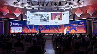Konferenzsaal mit Publikum, Bühne mit zwei Rednern, großer Bildschirm zeigt Präsentation zum Thema Energieversorgung und Transport, Banner mit Text 'sicher vorangehen' und 'dena ENERGIEWENDE KONGRESS 2025'.