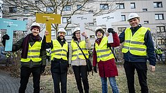 Fünf Personen in gelben Warnwesten und Helmen stehen im Freien. Sie halten bunte Schilder mit den Aufschriften 'energie de' und 'energie strong'.