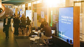Innenraum mit mehreren Personen auf einem Event oder einer Messe, Stehtische und große Bildschirme mit Texten an den Wänden