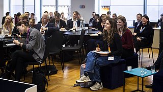 Mehrere Personen sitzen in einem hellen Raum mit großen Fenstern, einige schreiben in Notizbücher, andere hören zu.