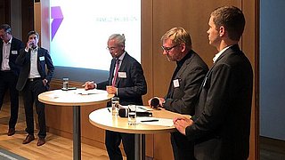 Mehrere Personen stehen vor einer Leinwand, Herr Joest hält ein Mikro