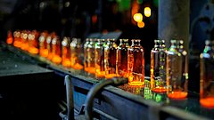 Reihe von transparenten Glasflaschen mit orangefarbenem Inhalt auf einem Förderband in einer Fabrik. Unscharfer Hintergrund mit grünen und gelben Lichtern.