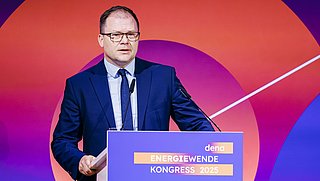 Carsten Schneider, Bundesminister für Umwelt, Klimaschutz, Naturschutz und nukleare Sicherheit, in blauem Anzug und Krawatte steht hinter einem Rednerpult mit dem Schriftzug 'dena ENERGIEWENDE KONGRESS 2025 sicher vorangehen' vor einem orange-violetten Hintergrund mit Kreisgrafik.