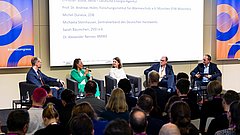 Im Vordergrund ist das Publikum zu sehen, im Hintergrund die Bühne mit den Diskutierenden. 