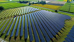 Luftaufnahme eines Solarparks mit mehreren Reihen von Solarpanelen auf einer grünen Wiese, umgeben von Feldern in verschiedenen Farben.