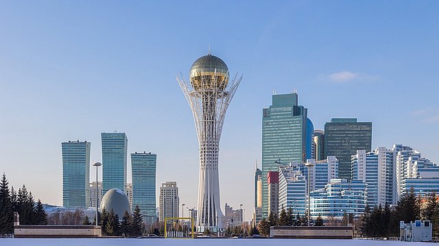 Stadtansicht von Astana mit dem Bayterek-Turm im Vordergrund, umgeben von modernen Hochhäusern und Bäumen.