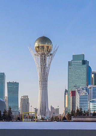 Stadtansicht von Astana mit dem Bayterek-Turm im Vordergrund, umgeben von modernen Hochhäusern und Bäumen.