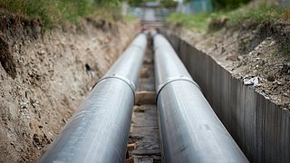 Zwei große Metallrohre liegen parallel in einem Erdgraben, umgeben von Erde und Vegetation.
