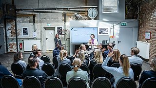 Zwei Personen halten einen Vortrag vor einem Publikum. Menschen im Publikum melden sich.