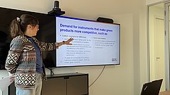 Frau mit Zopf und gemustertem Pullover zeigt auf Bildschirm mit englischem Text zu grünen Produkten in einem Besprechungsraum.