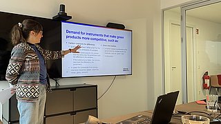 Frau mit Zopf und gemustertem Pullover zeigt auf Bildschirm mit englischem Text zu grünen Produkten in einem Besprechungsraum.