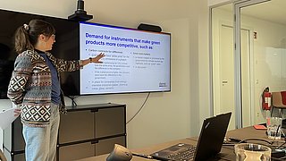 Frau mit Zopf und gemustertem Pullover zeigt auf Bildschirm mit englischem Text zu grünen Produkten in einem Besprechungsraum.