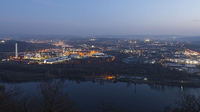 Nächtliche Panoramaaufnahme einer Industrieanlage und Stadt, beleuchtet mit Straßenlichtern, neben einem Fluss.