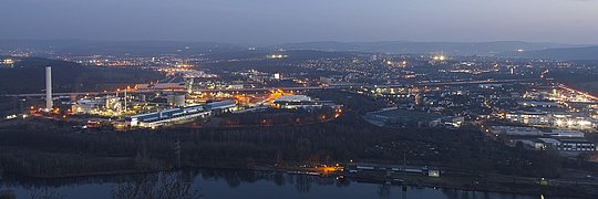 Nächtliche Panoramaaufnahme einer Industrieanlage und Stadt, beleuchtet mit Straßenlichtern, neben einem Fluss.