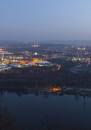 Nächtliche Panoramaaufnahme einer Industrieanlage und Stadt, beleuchtet mit Straßenlichtern, neben einem Fluss.