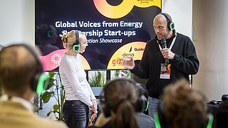 Mehrere Personen mit Kopfhörern sitzen und stehen vor einem großen Bildschirm mit Veranstaltungstitel.