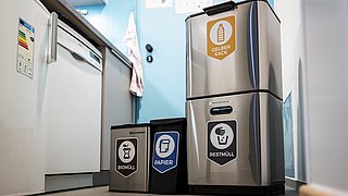 Drei Müllbehälter in einer Küche: ein grauer Behälter für 'RESTMÜLL', ein schwarzer für 'PAPIER' und ein blauer für 'BIOMULL'. Der graue Behälter hat eine doppelte Funktion.