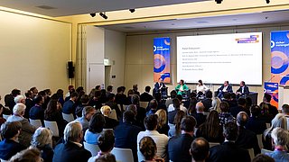 Fünf Personen auf der Bühne diskutieren, hinter ihnen eine Powerpointfolie auf der die Namen der Personen gelistet sind. 