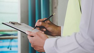 Nahaufnahme von einem Oberkörper und Händen, die ein Klemmbrett mit einem Formular halten. Mit einem Stift schreibt die Person ins Formular.