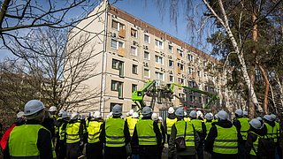 Gruppe von Personen mit Schutzhelmen und Warnwesten vor einem mehrstöckigen Wohngebäude mit teilweise mit Holzplatten verschlossenen Fenstern und zwei Arbeitsbühnen an der Fassade