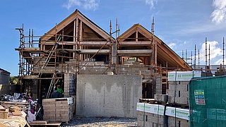 Haus im Rohbau mit Holzgiebeln und Gerüst, Baumaterialien und Bauzaun im Vordergrund, blauer Himmel mit Wolken