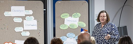 Hanne May, Bereichsleiterin Kommunikation, steht vor Pinnwänden mit beschrifteten Zetteln und spricht zu einer Gruppe von Zuhörern, die ihr Rücken zugewandt sind.