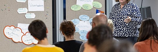 Hanne May, Bereichsleiterin Kommunikation, steht vor Pinnwänden mit beschrifteten Zetteln und spricht zu einer Gruppe von Zuhörern, die ihr Rücken zugewandt sind.