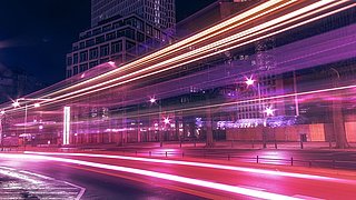 Lichtspuren von Fahrzeugen auf einer Straße vor Hochhäusern bei Nacht