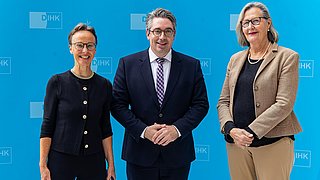 Drei Personen stehen vor einem blauen Hintergrund mit dem Text 'DIHK'. Die linke Person trägt einen schwarzen Pullover mit Knöpfen, die mittlere Person trägt einen Anzug mit Krawatte, die rechte Person trägt einen beigen Blazer.