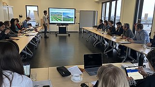 Konferenzraum mit mehreren Personen an Tischen, eine Person steht vor Bildschirm mit Präsentation zu Landwirtschaft.