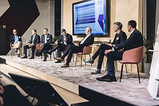 Sechs Personen sitzen auf einem Podium, mit Mikrofonen und Getränken. Im Hintergrund ein Bildschirm mit Grafiken und Text: 'Wie der Rollout gelingt'.