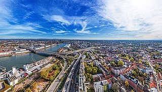 Panoramaaufnahme einer Stadt mit Fluss, Brücken, Straßen und bunten Gebäuden unter blauem Himmel.