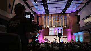 Kameramann filmt Vortrag in großem Saal mit Orgel und Publikum
