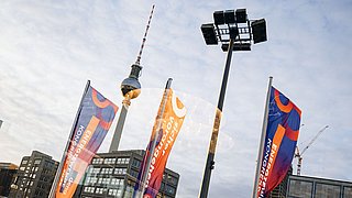 Mehrere bunte Fahnen vor dem Berliner Fernsehturm und umliegenden Gebäuden bei bewölktem Himmel