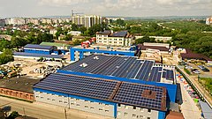 Luftaufnahme eines großen Gebäudes mit blauer Fassade und einem Dach, das mit Solarpanelen bedeckt ist. Im Hintergrund sind mehrere Wohngebäude und eine grüne Landschaft zu sehen.