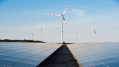 Windkraftanlagen stehen hinter Solarpanelen auf einem Feld, unter blauem Himmel mit wenigen Wolken.