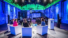 Vier Personen sitzen auf einer Bühne in weißen Sesseln und diskutieren vor Publikum in einem bunt beleuchteten, industriellen Veranstaltungsraum.