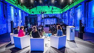 Vier Personen sitzen auf einer Bühne in weißen Sesseln und diskutieren vor Publikum in einem bunt beleuchteten, industriellen Veranstaltungsraum.