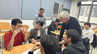 Gruppe von vietnamesischen Studierenden sitzen in einem Seminarraum an Tischen. Ein Dozent steht neben ihnen. Sie diskutieren zusammen.