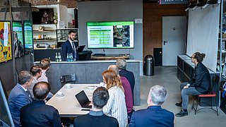 Zu sehen sind Personen, die eine Präsentation halten und Menschen an einem Gruppentisch