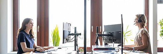 Zwei Frauen sitzen an Schreibtischen vor einem Fenster und arbeiten konzentriert an ihren Aufgaben.