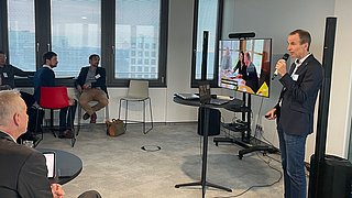 Mann mit Mikrofon steht vor Bildschirm, auf dem zwei Frauen zu sehen sind, mehrere Personen sitzen im Raum mit großen Fenstern.